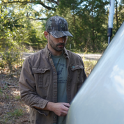 Man wearing a Mossy oak camouflage cap and brown jacket standing outdoors with trees in the background