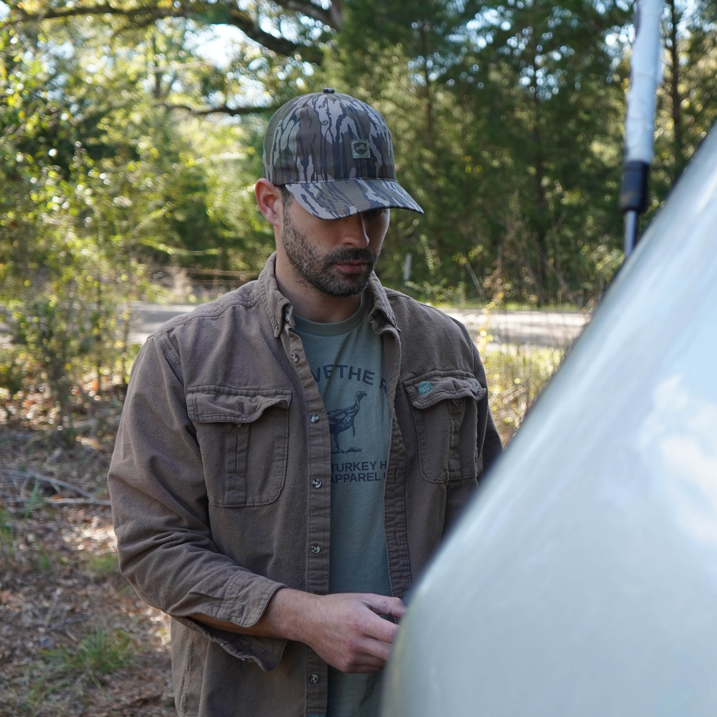 Man wearing a Mossy oak camouflage cap and brown jacket standing outdoors with trees in the background